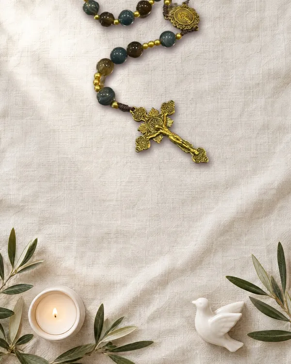 Terço Azul e marrom com crucifixo dourado, perto de uma vela acesa, ramos de oliveira e uma pomba de cerâmica
